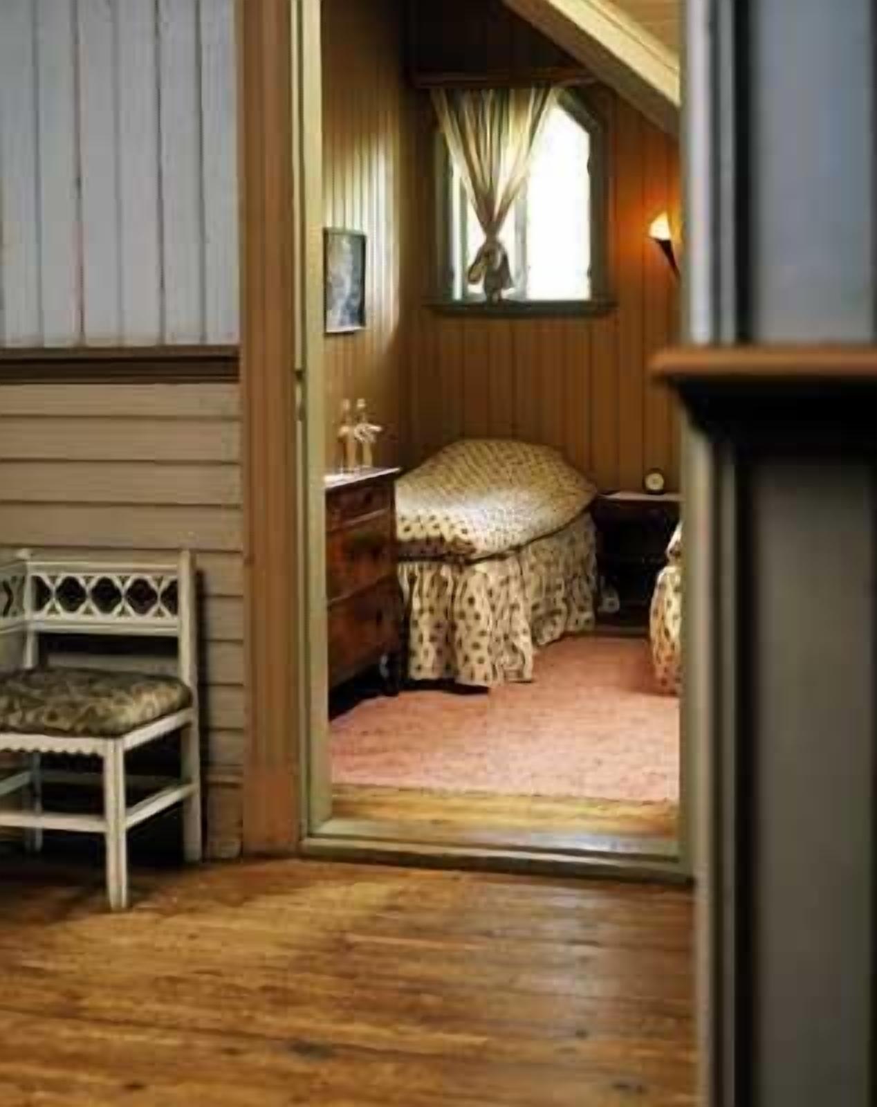 A cozy bedroom with a wooden floor, a bed with a patterned bedspread, a nightstand, a lamp, and a bench.