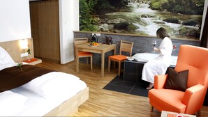 A hotel room with a bed, a dining table with chairs, a small kitchen area, and a large window with a view of a waterfall. - Almfrieden (Ramsau am Dachstein)