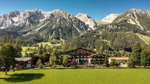 A mountain lodge with multiple floors, surrounded by a lush green field and forested mountains. - Almfrieden (Ramsau am Dachstein)