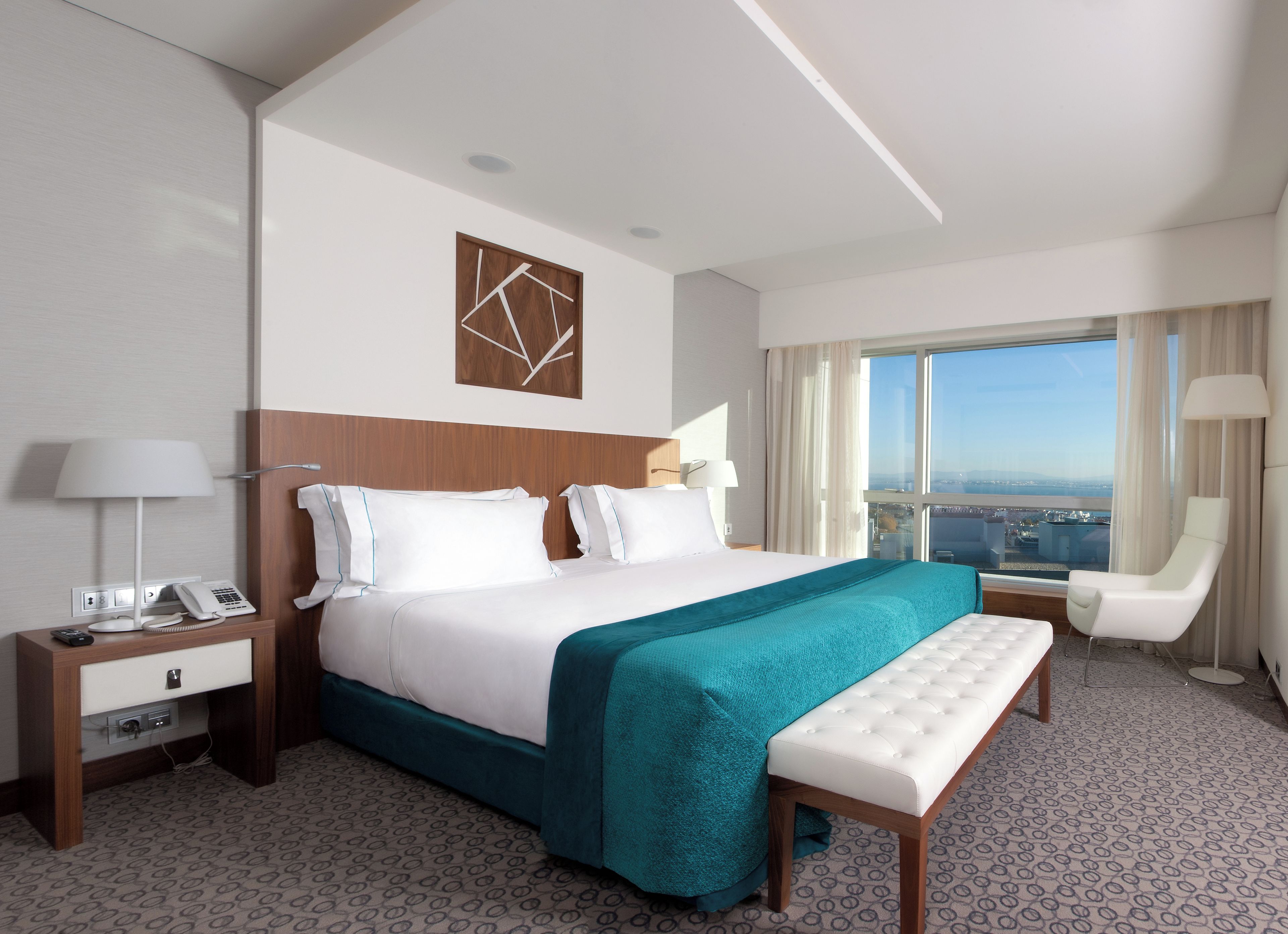 Ein Hotelzimmer mit einem großen Bett, einem Nachttisch mit Lampe, einem Sessel und Blick auf die Stadt.