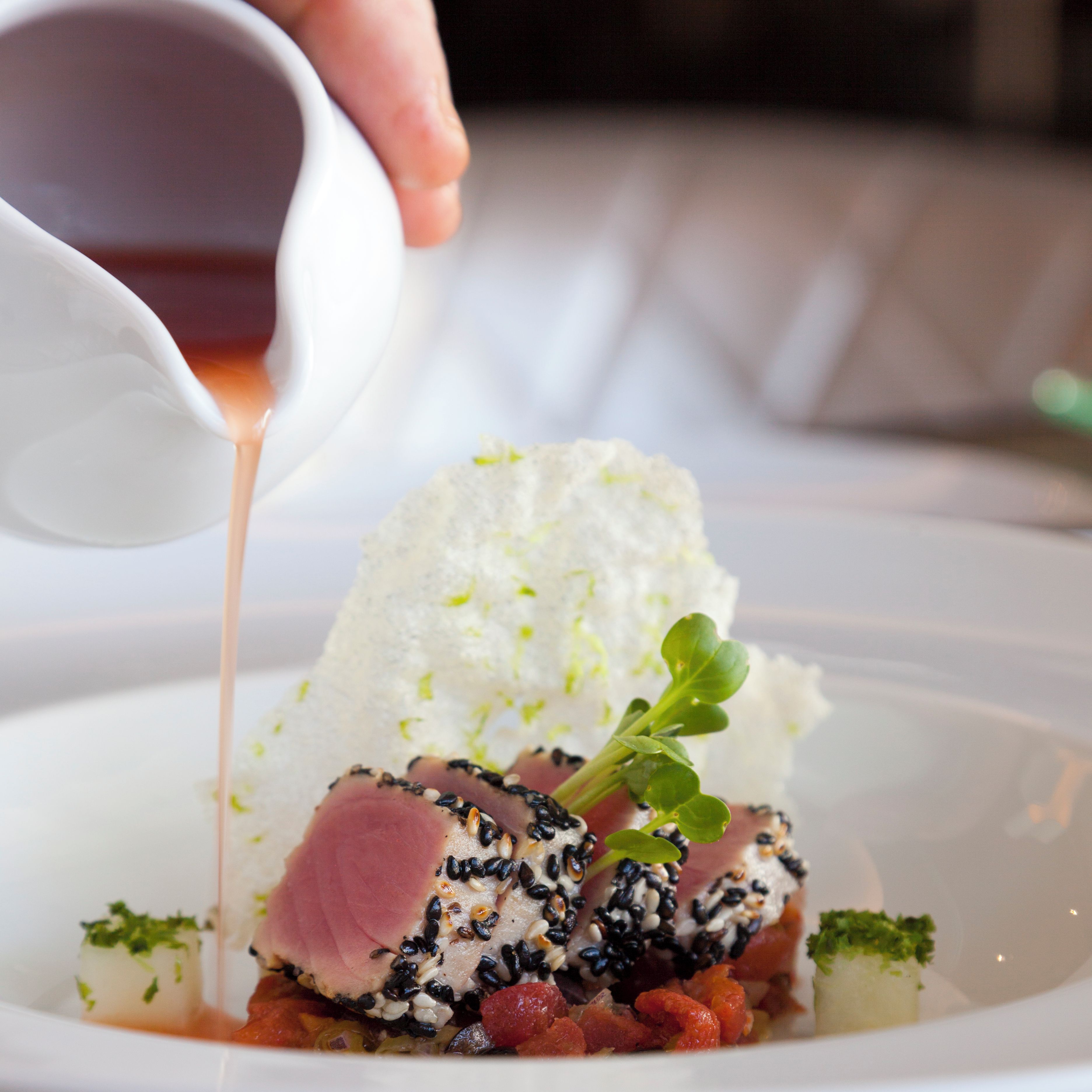 Une assiette blanche contenant un plat composé d’une tranche de viande recouverte de graines de sésame noir, d’une sauce blanche crémeuse et décorée de feuilles vertes.