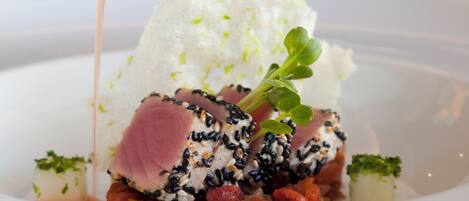 A white plate with a dish consisting of a slice of meat topped with black sesame seeds, a white creamy substance, and garnished with green leaves.