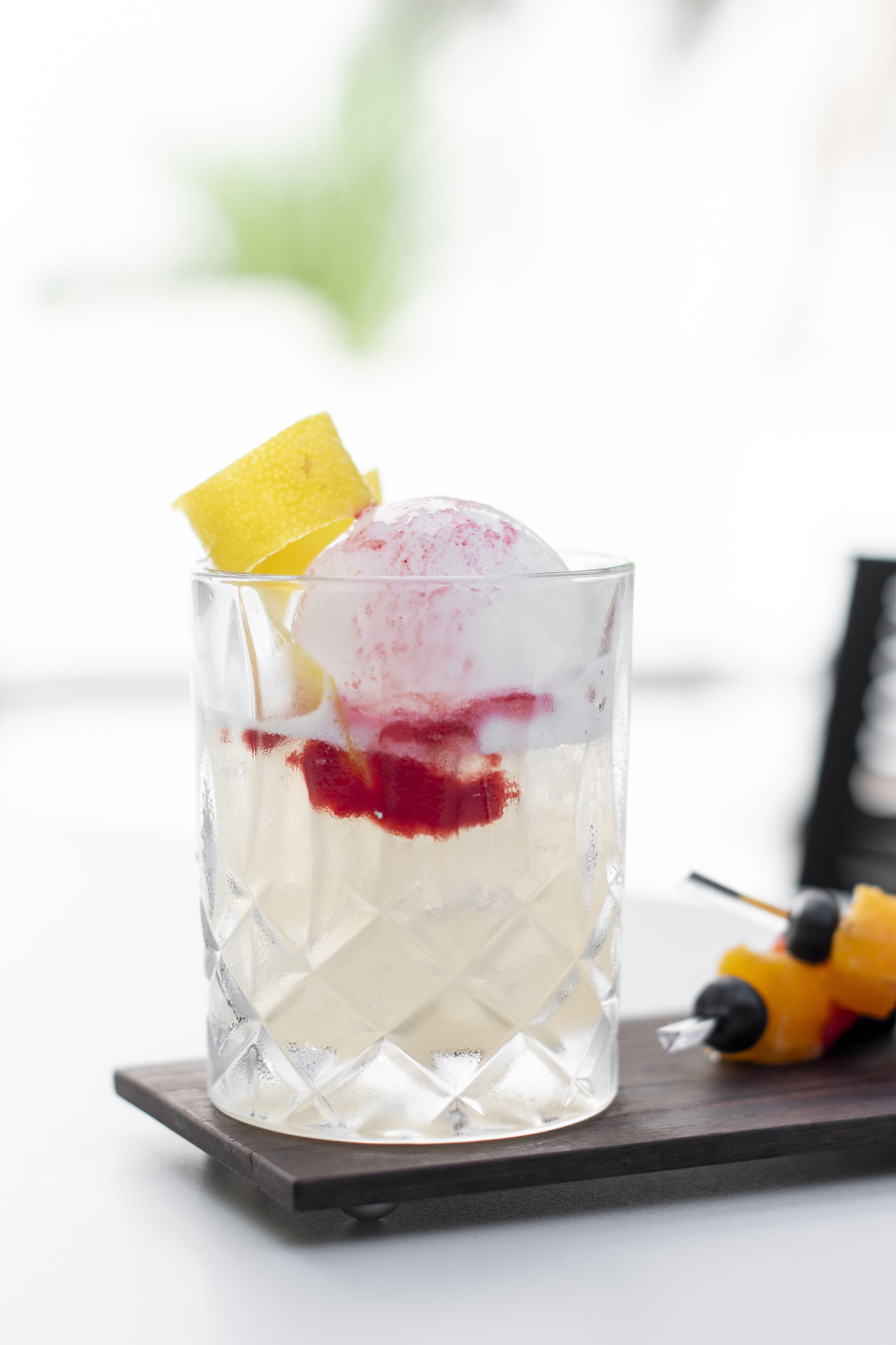 A glass with a layered dessert, a slice of lemon, and a yellow fruit on a wooden coaster.