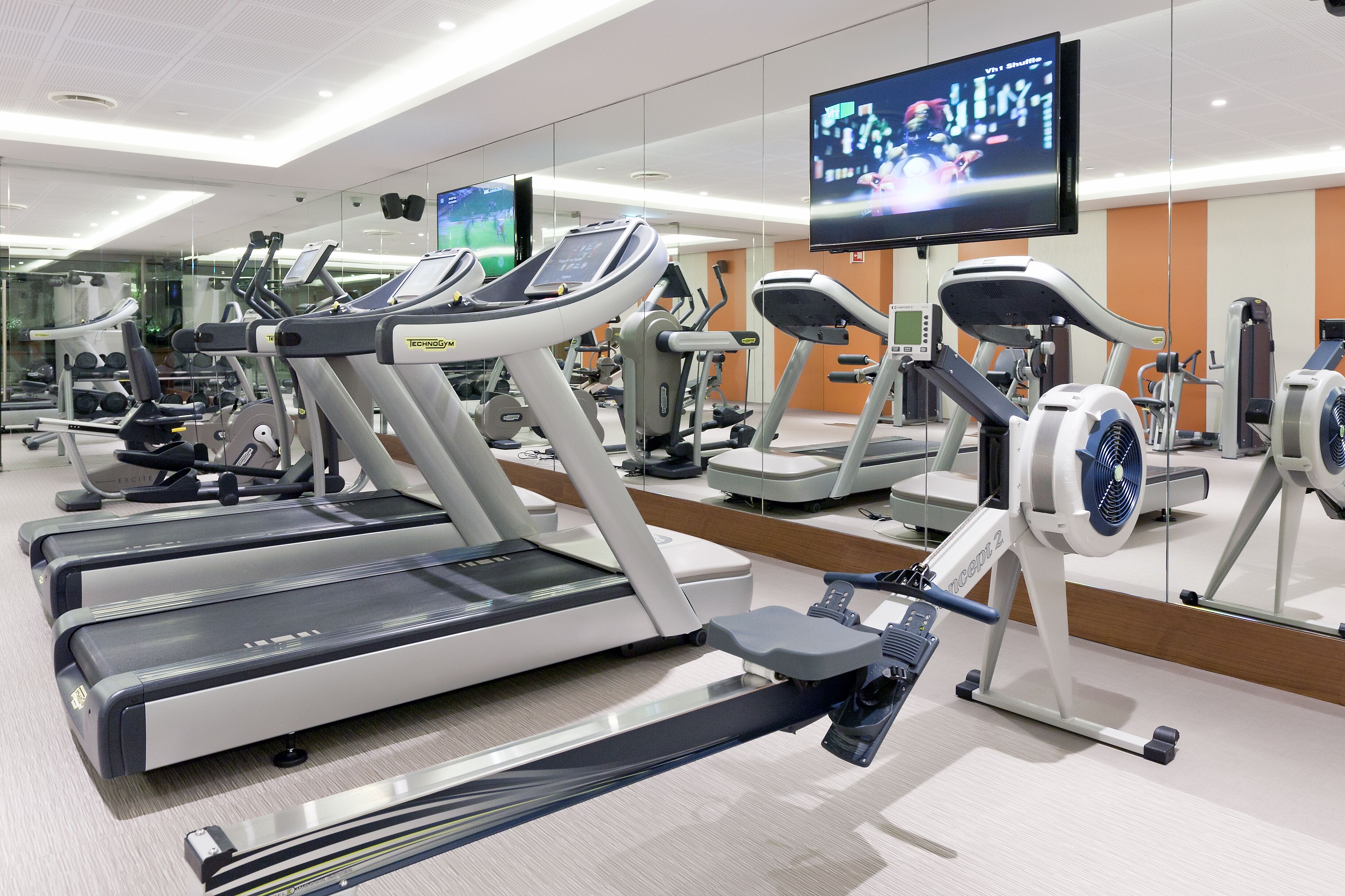 Une salle de sport équipée de plusieurs tapis de course, de vélos d’exercice et d’un écran de télévision diffusant un événement sportif.