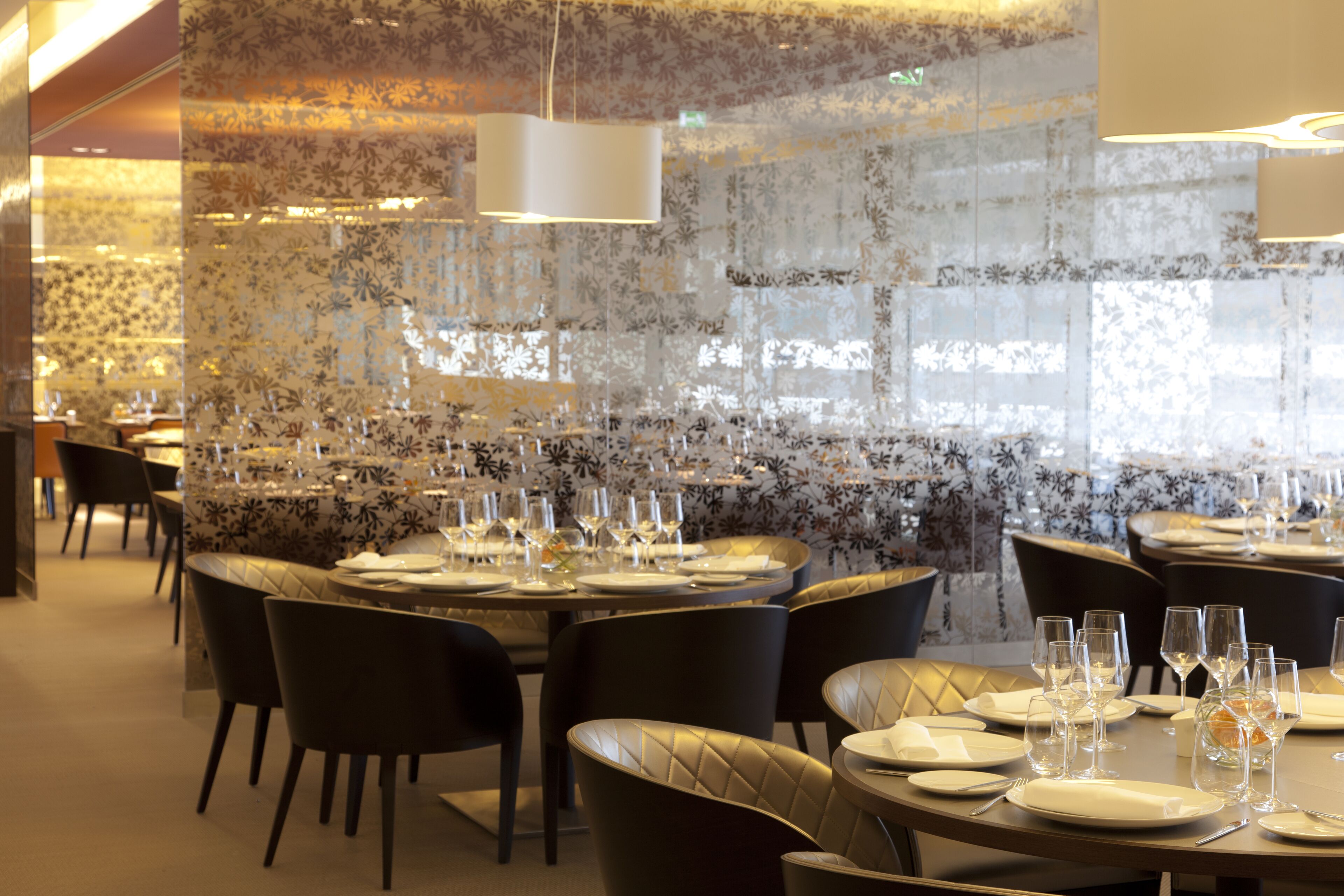 Restaurante com mesas redondas preparadas para refeições, apresentando uma parede estampada e iluminação moderna.