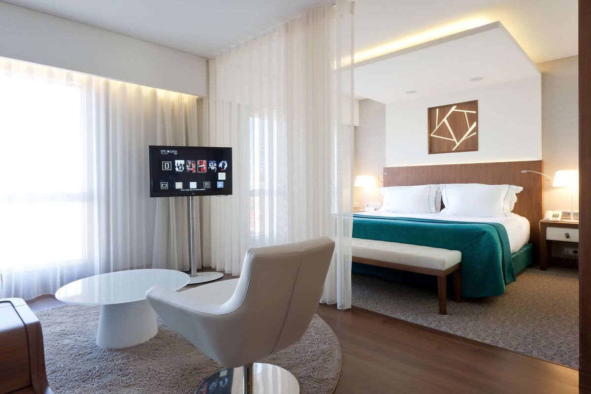 A modern hotel room with a bed, a TV, a white armchair, and a small table.