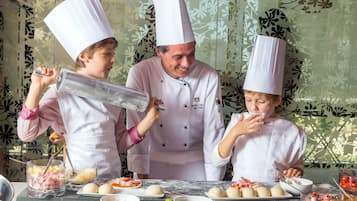 Três pessoas com chapéus de chef preparando comida com diversos ingredientes e utensílios sobre uma mesa.