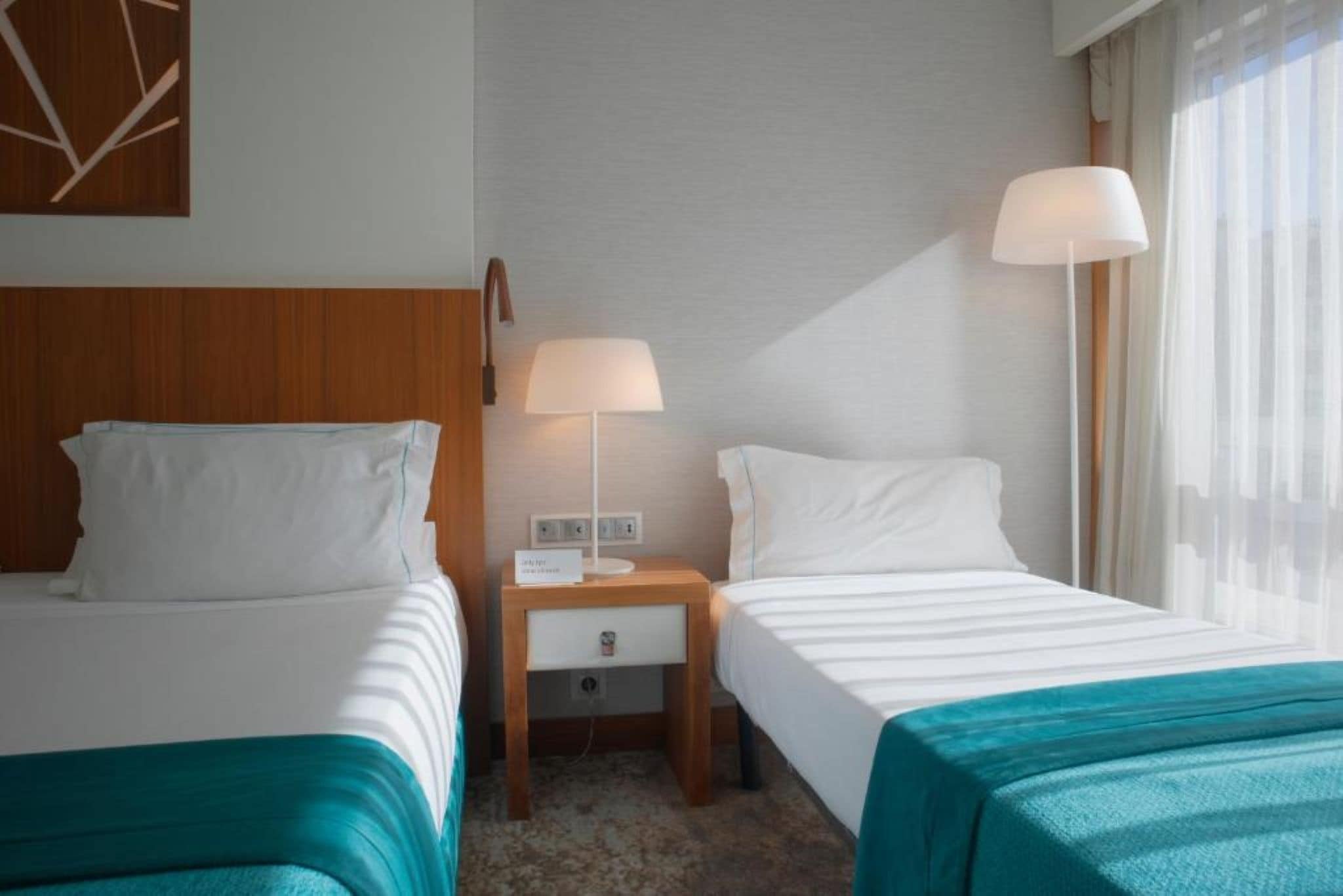 A hotel room with two beds, a nightstand with a lamp, and a window with curtains.