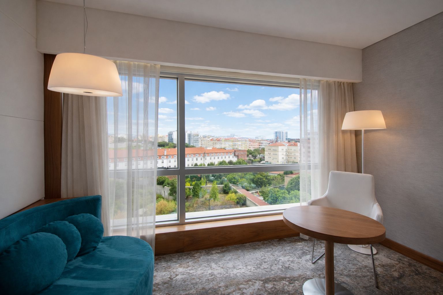 Habitación de hotel con un ventanal grande, un sofá azul, una silla blanca y una mesita.