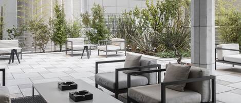 A modern outdoor seating area with white sofas and a table, surrounded by greenery and a large window.