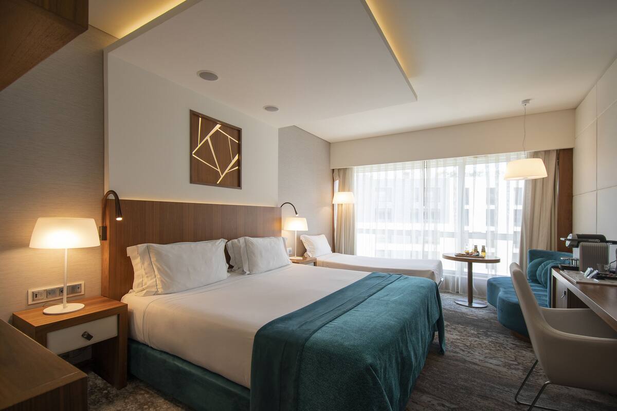 A hotel room with a large bed, two bedside tables with lamps, a desk with a chair, and a view of the city through large windows.