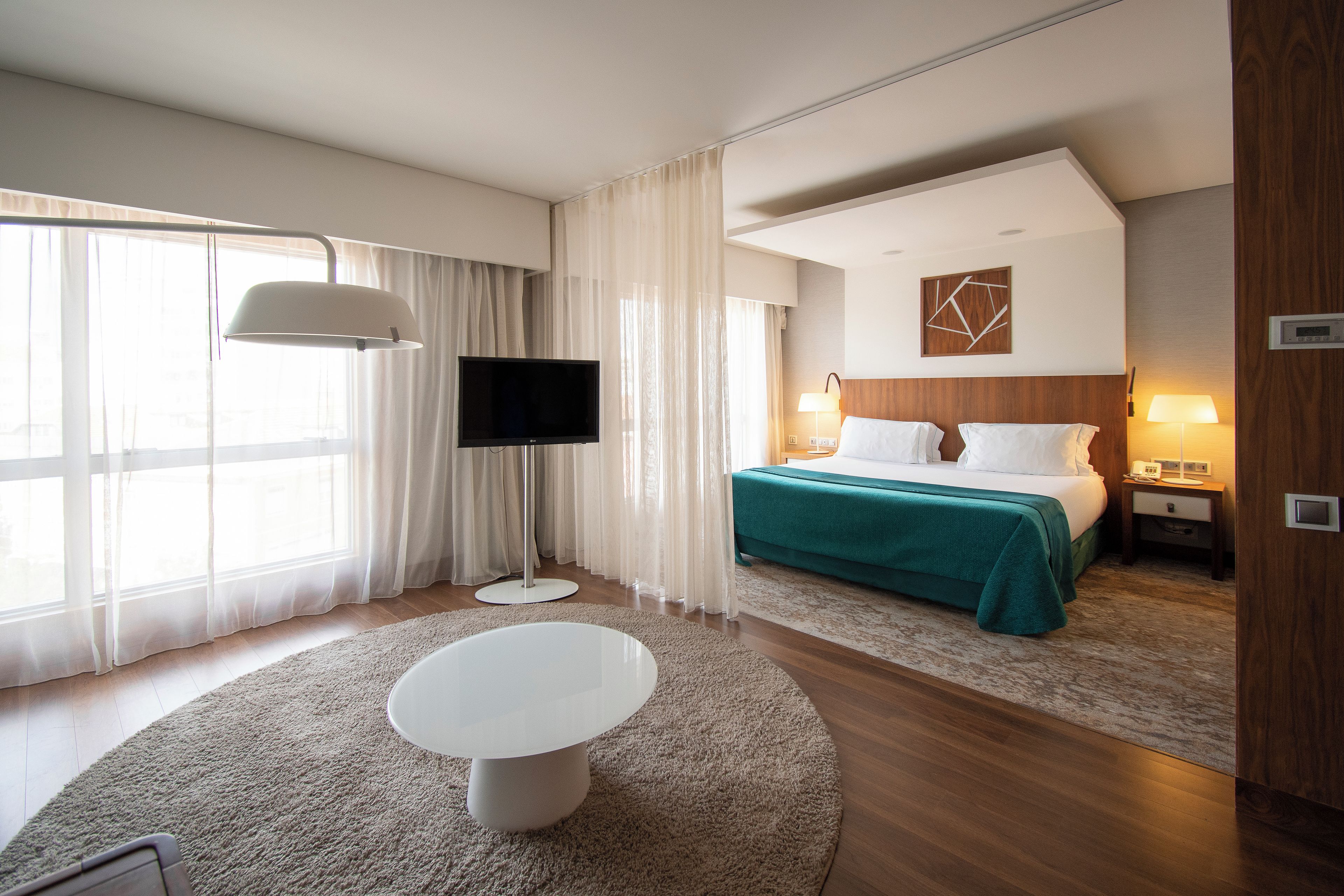 A hotel room with a bed, a TV, a desk, and a window with curtains.