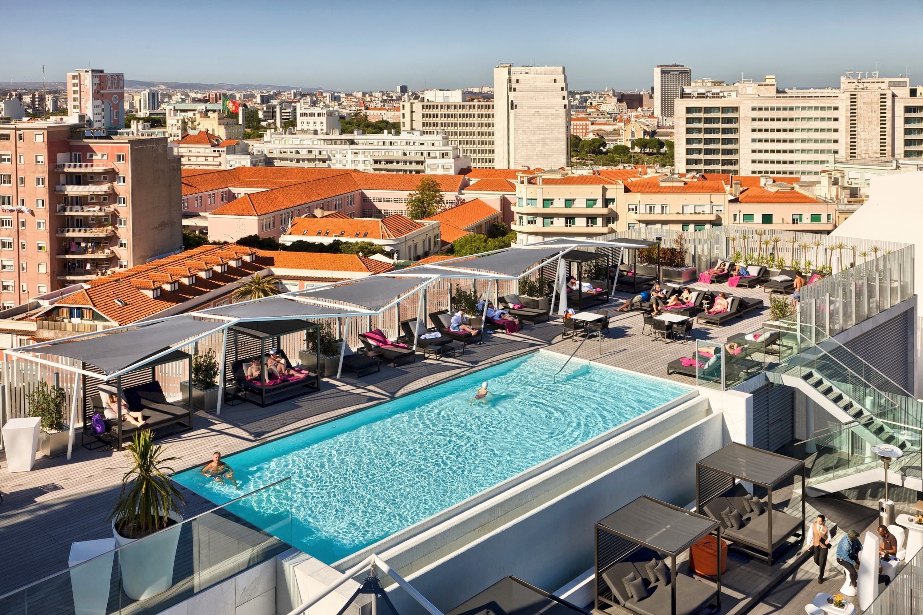 A rooftop pool with lounge areas, a glass balustrade, and a cityscape view.