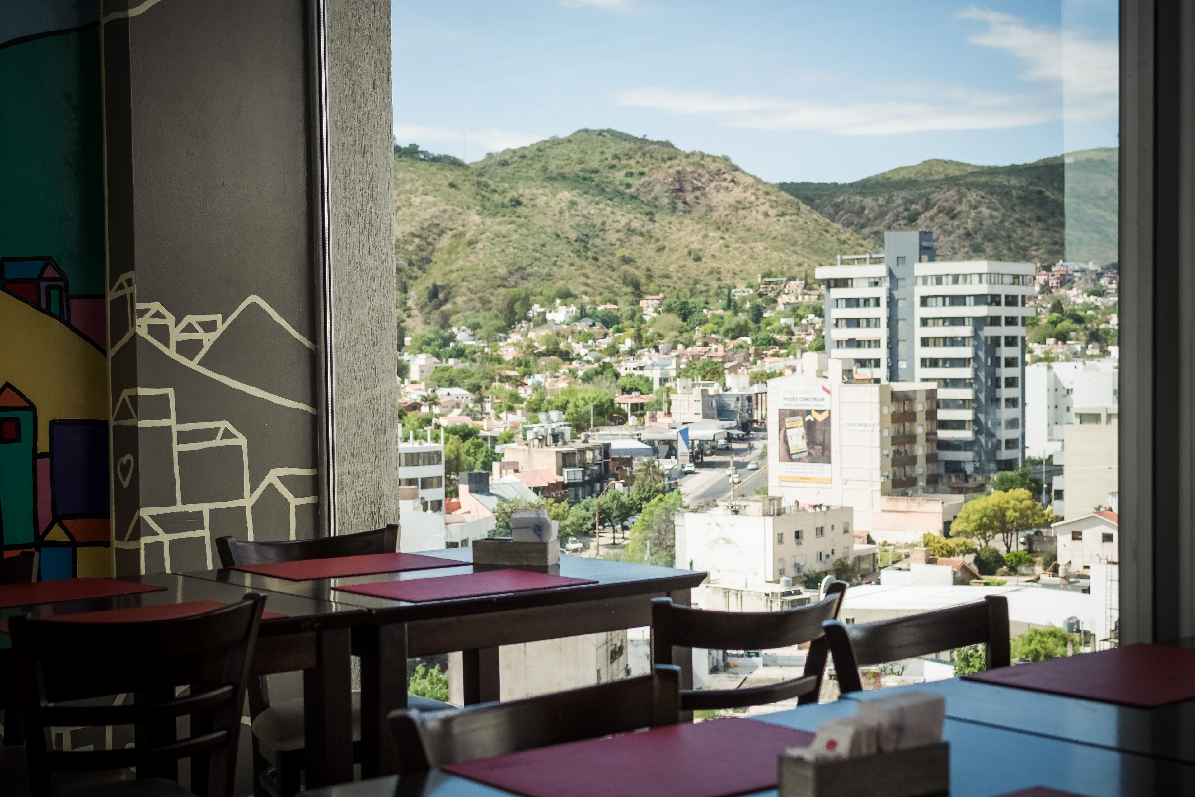Un restaurante con mesas y sillas, con vista a un paisaje urbano que incluye edificios y colinas al fondo.