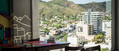 Un restaurante con mesas y sillas, con vista a un paisaje urbano que incluye edificios y colinas al fondo.