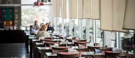 Un restaurante espacioso con mesas y sillas largas, ventanas amplias y lámparas rojas colgantes.