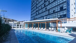 Una piscina con sillones y mesas, rodeada por un edificio moderno con grandes ventanales.
