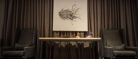 A room with a dark curtain backdrop, a framed artwork on a table, two armchairs, and a small statue.