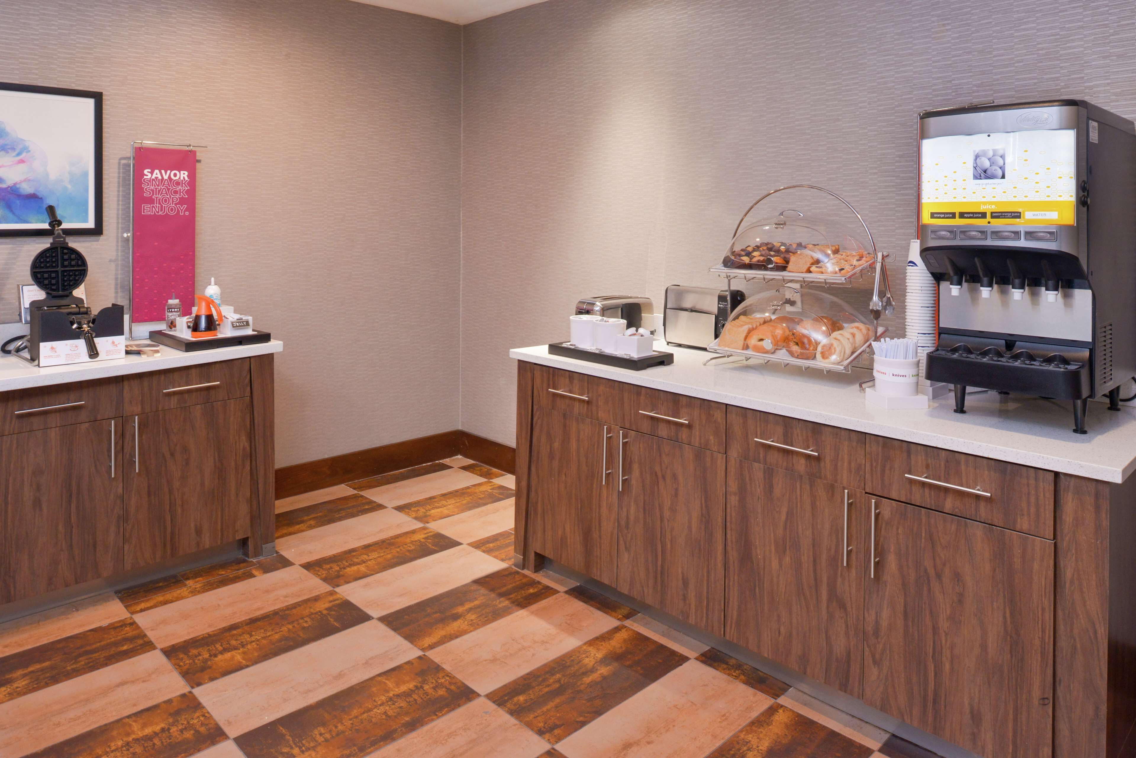 A hotel room with a checkered floor, wooden cabinets, a coffee machine, and a breakfast bar with pastries.
