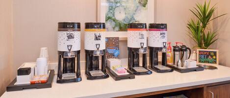 A coffee station with multiple coffee makers, a welcome sign, and a plant.