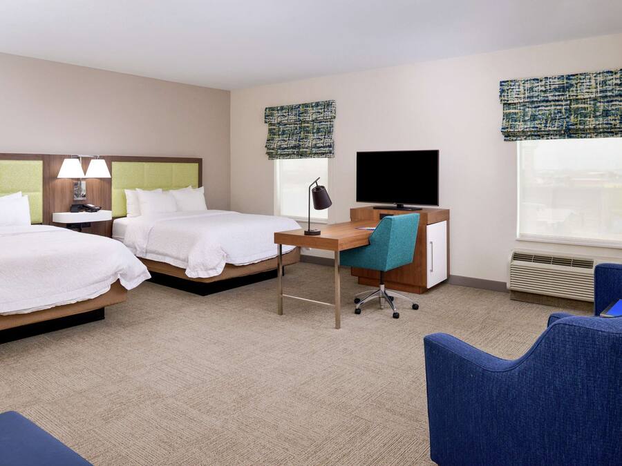 A hotel room with two beds, a desk with a chair, a television, and a large window with blinds.