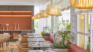 A restaurant with long communal tables, cushioned seating, hanging pendant lights, and large windows with views of greenery. - Zafiro Ca'n Picafort (Santa Margalida)