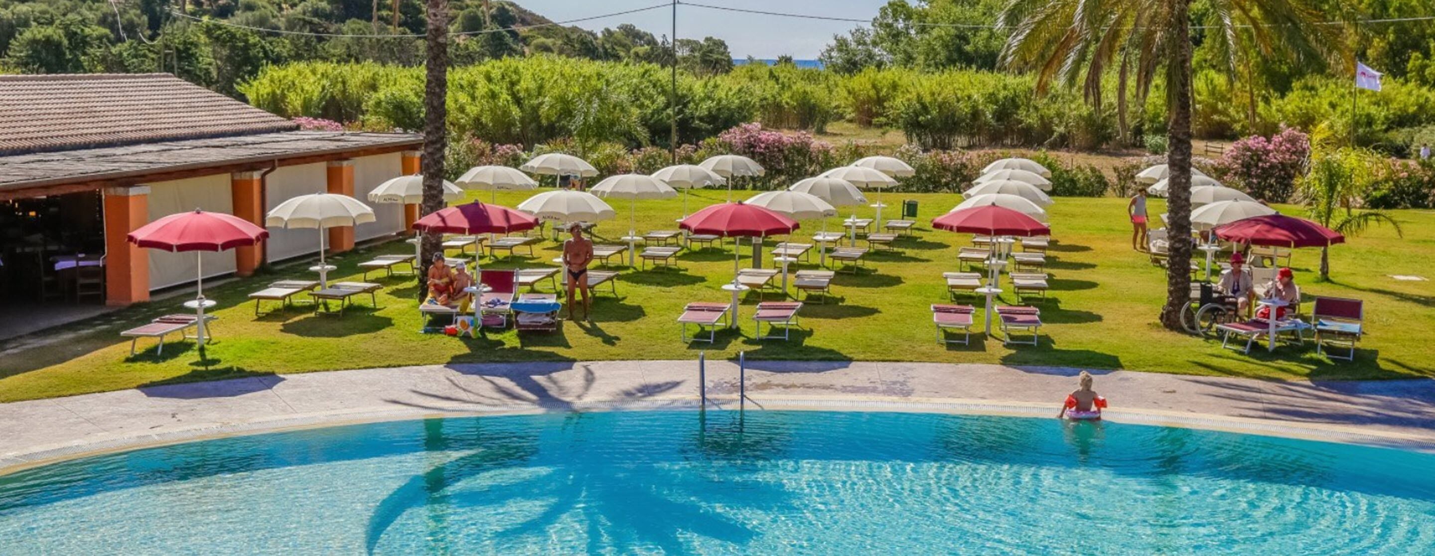 A pool area with multiple umbrellas, lounge chairs, and a building with a tiled roof.