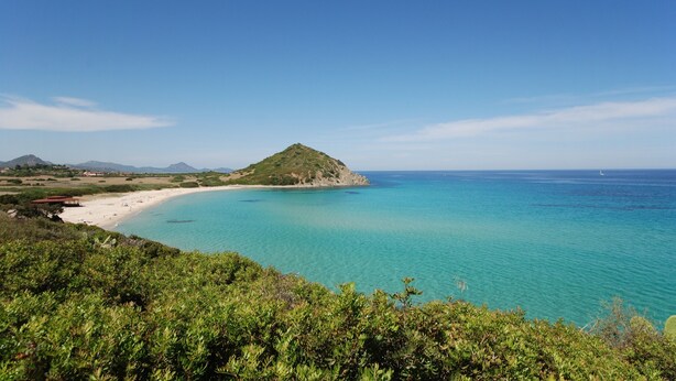 A coastal landscape with a beach, clear turquoise waters, and a hill with vegetation.