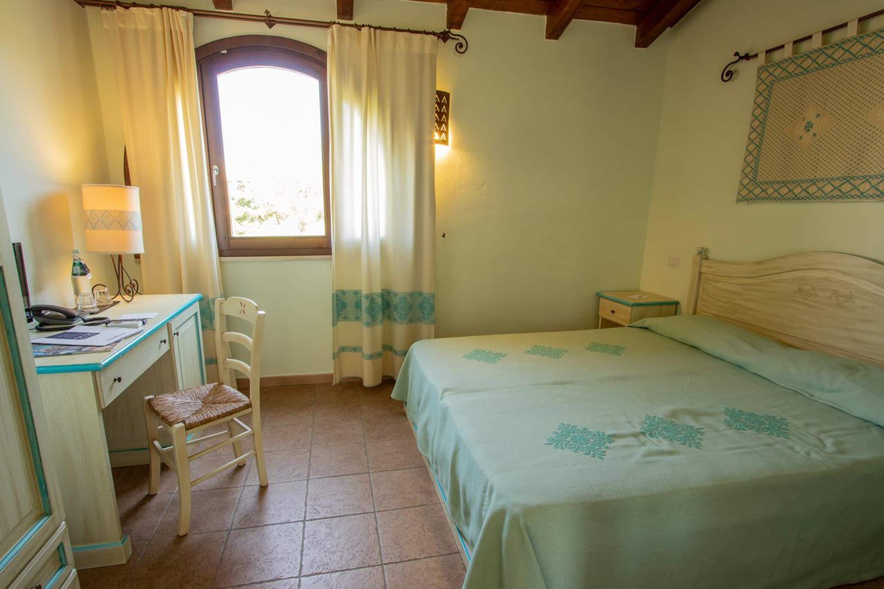 Ein Schlafzimmer mit Bett, Schreibtisch, Stuhl, einem Fenster mit Vorhängen, einem Nachttisch und einer Wandlampe.