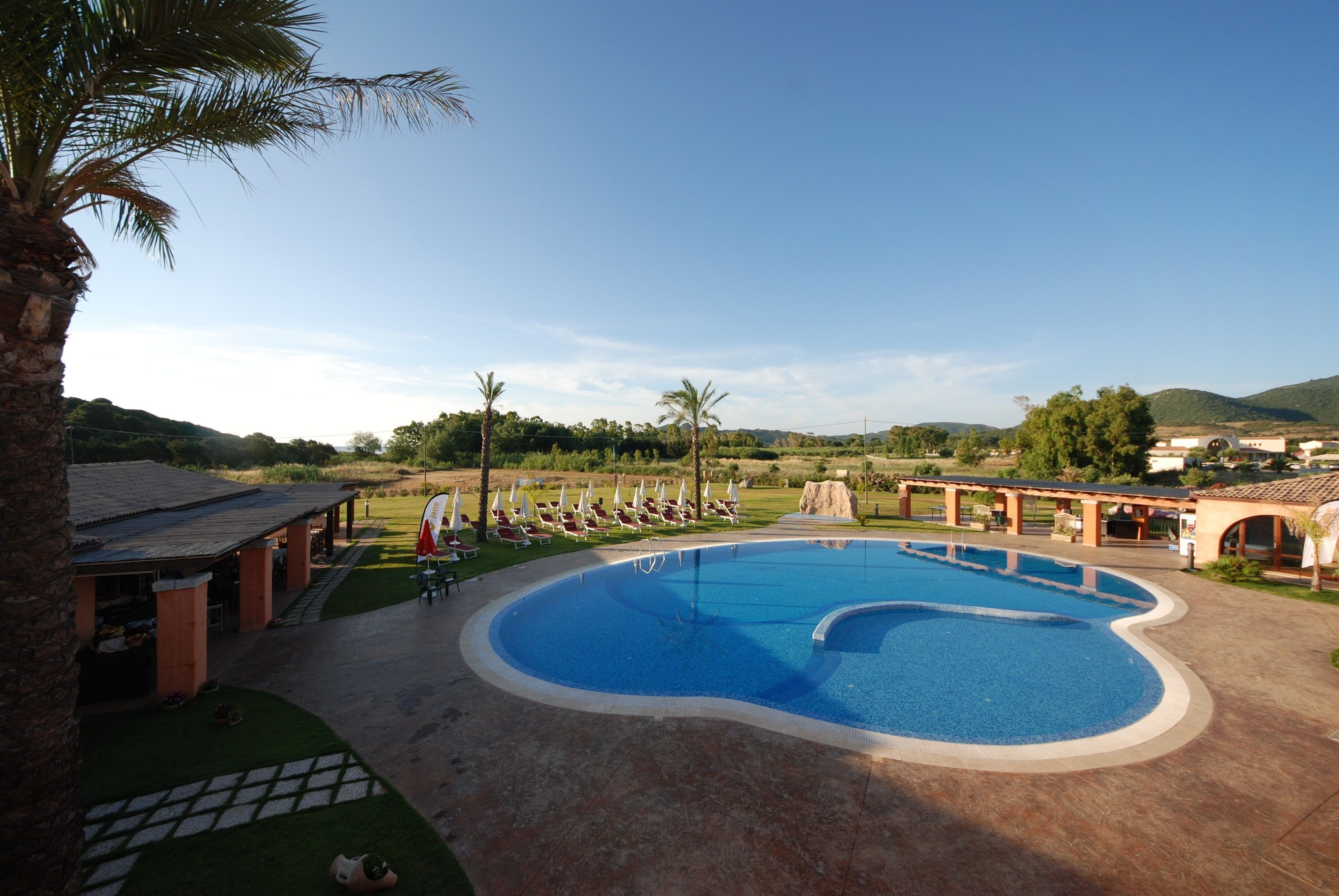 A swimming pool with lounge chairs and umbrellas, surrounded by palm trees and a building with a tiled roof.