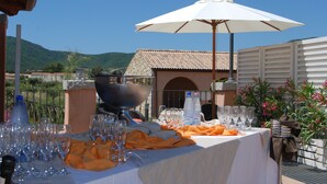 An outdoor dining area with a table set for a meal, surrounded by a scenic view of hills and a building with a tiled roof.
