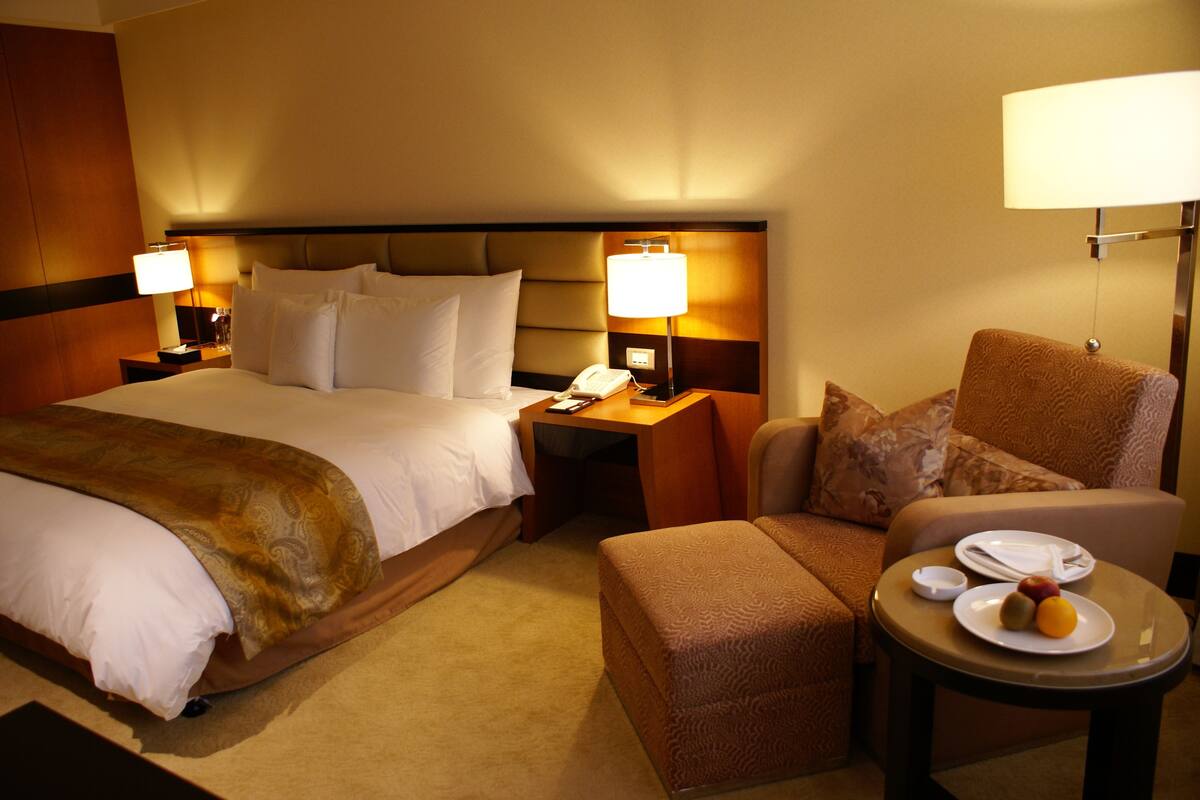 Una habitación de hotel con una cama grande, dos mesitas de noche, una silla, una mesa pequeña con un plato de fruta y una lámpara en cada mesita de noche.