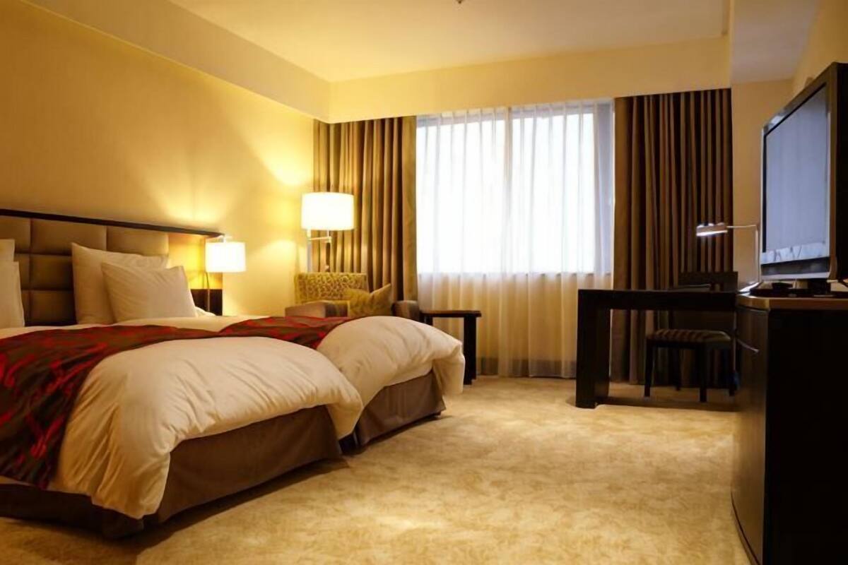 Una habitación de hotel con una cama grande, un escritorio con computadora, televisor y ventana con cortinas.