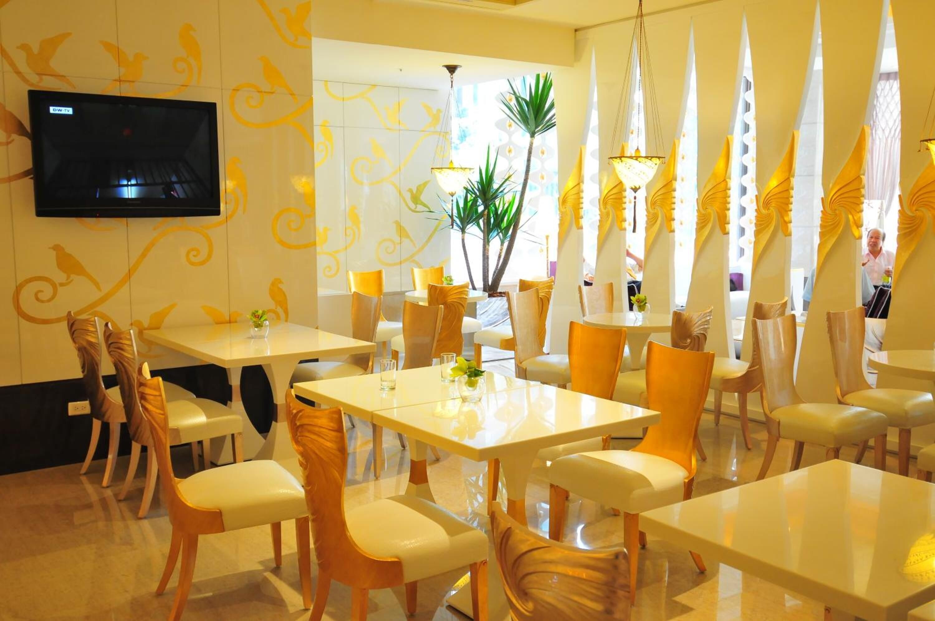 Un espace repas avec des tables et des chaises blanches, un téléviseur fixé au mur et des motifs décoratifs sur les murs.