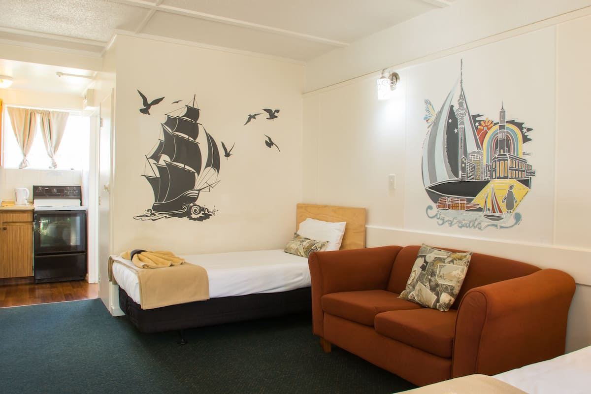 Quarto com cama, sofá, pequeno espaço de cozinha e arte nas paredes retratando navios e uma paisagem urbana.
