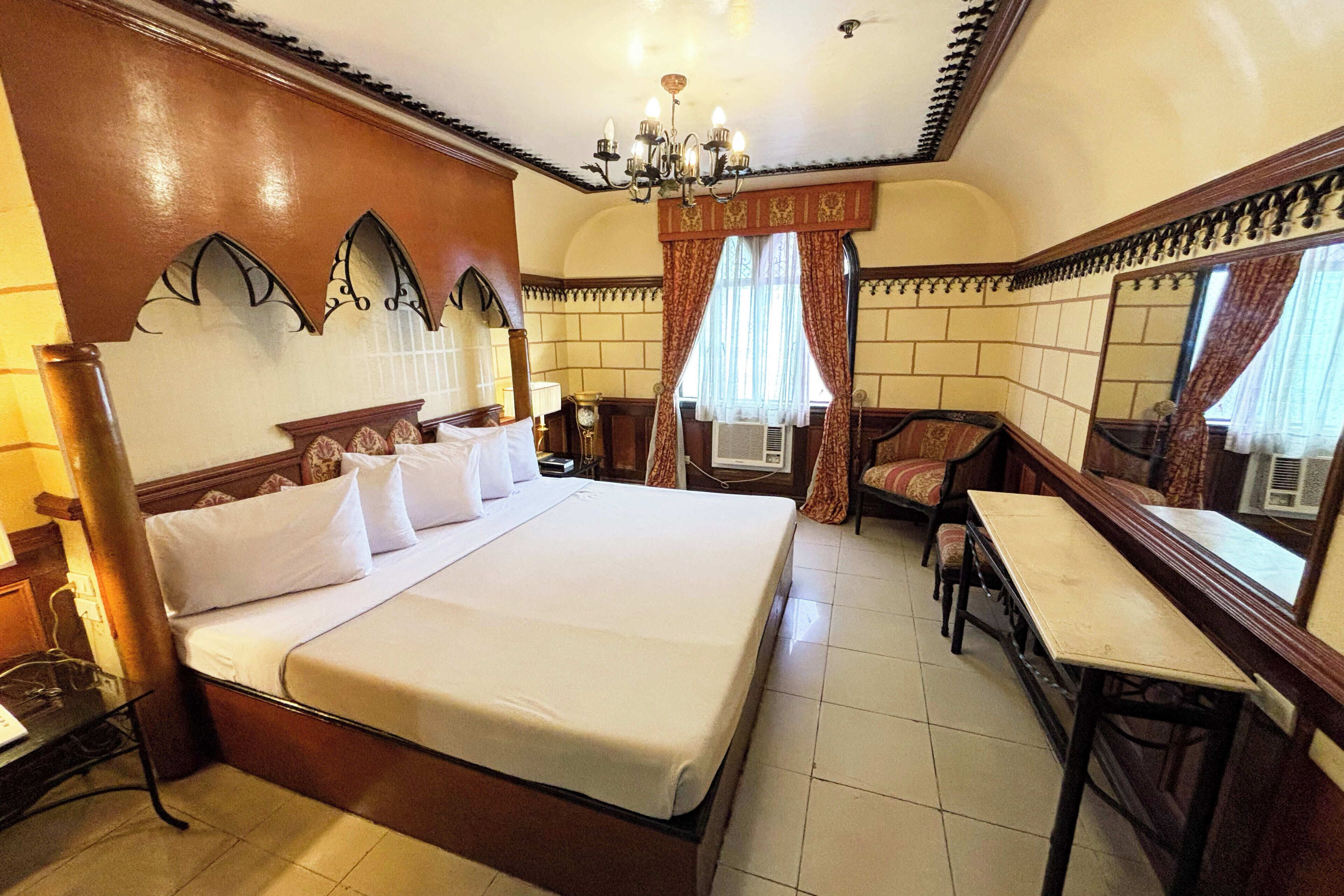 Une chambre d’hôtel avec un grand lit, un bureau et une chaise. On y trouve un lustre, des rideaux et un miroir au mur.