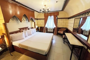 A hotel room with a large bed, a desk, and a chair. There is a chandelier, curtains, and a mirror on the wall. - Camelot Hotel Quezon City powered by Cocotel (Quezon City)