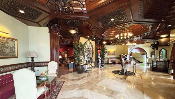 Un vaste hall d’entrée avec un sol en marbre, un plafond en bois richement décoré et un lustre. On y trouve des coins salon avec un canapé rouge et un fauteuil blanc.