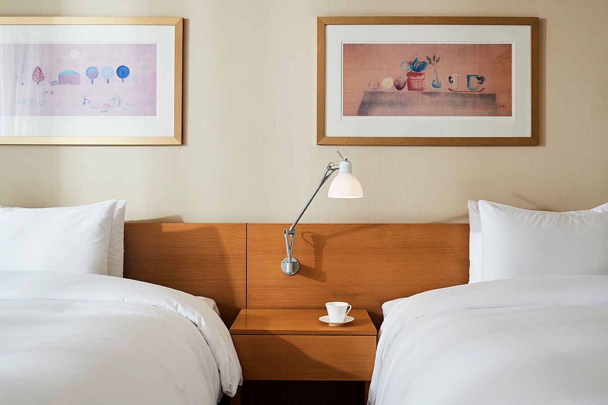 A hotel room with two beds, a nightstand with a lamp, and two framed artworks on the wall.