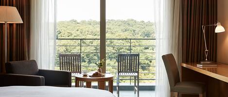 A hotel room with a bed, a desk, two chairs, a lamp, and a balcony with a view of greenery.