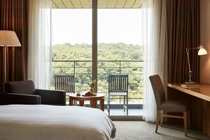 A hotel room with a bed, a desk, two chairs, a lamp, and a balcony with a view of greenery.