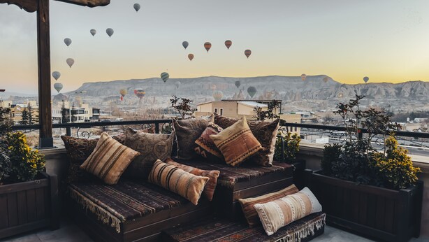 A balcony with a sofa and cushions, overlooking a cityscape with mountains in the background and hot air balloons in the sky. - Royal Stone Houses - Goreme (Nevsehir)