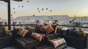 Un balcón con sofá y cojines, con vistas a un paisaje urbano con montañas al fondo y globos aerostáticos en el cielo.