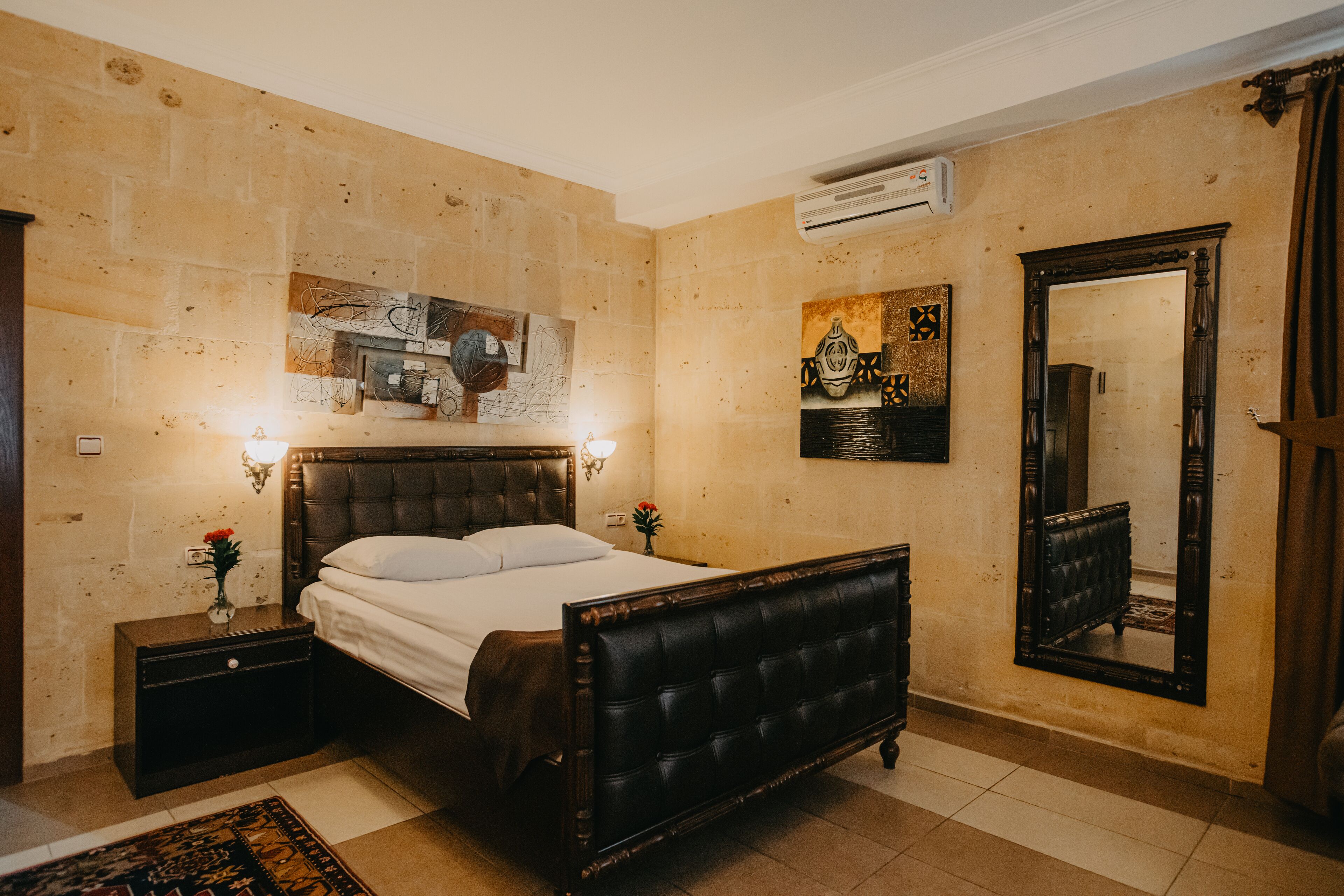 A bedroom with a large bed, a nightstand, a wall-mounted air conditioner, and a mirror.
