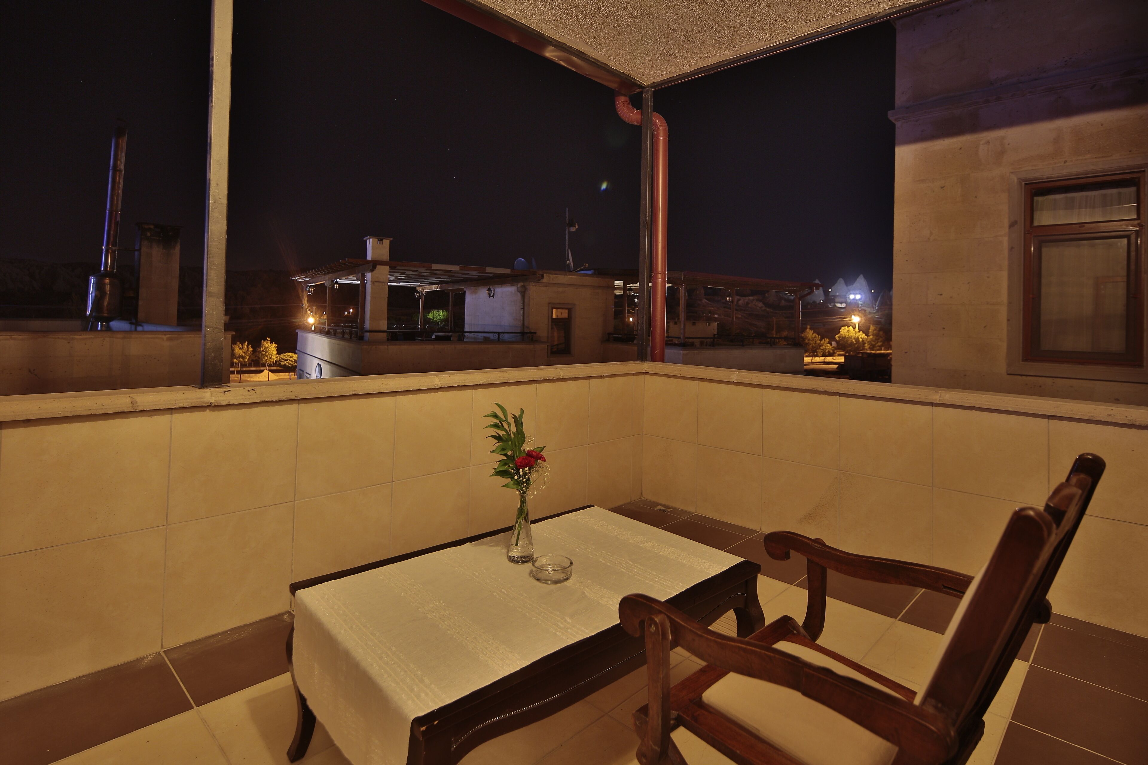 A balcony with a table and chairs, a vase with flowers, and a view of buildings at night.