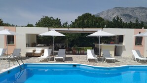 A pool area with lounge chairs and umbrellas, a bar, and a view of mountains. - Aphrodite Samos Superior Studios & Apartments (Samos)