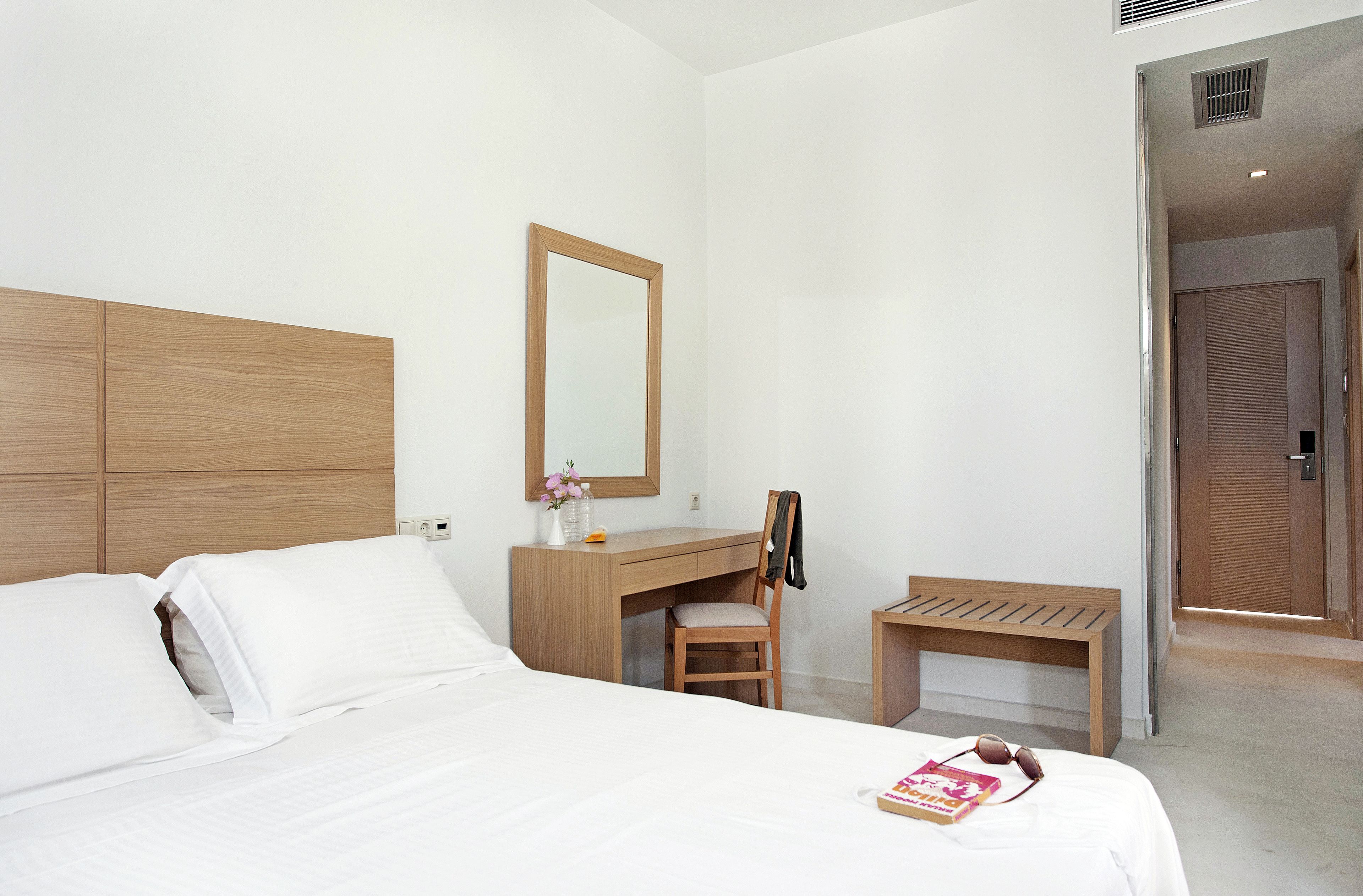 A hotel room with a bed, a wooden desk, a chair, a bench, and a mirror.