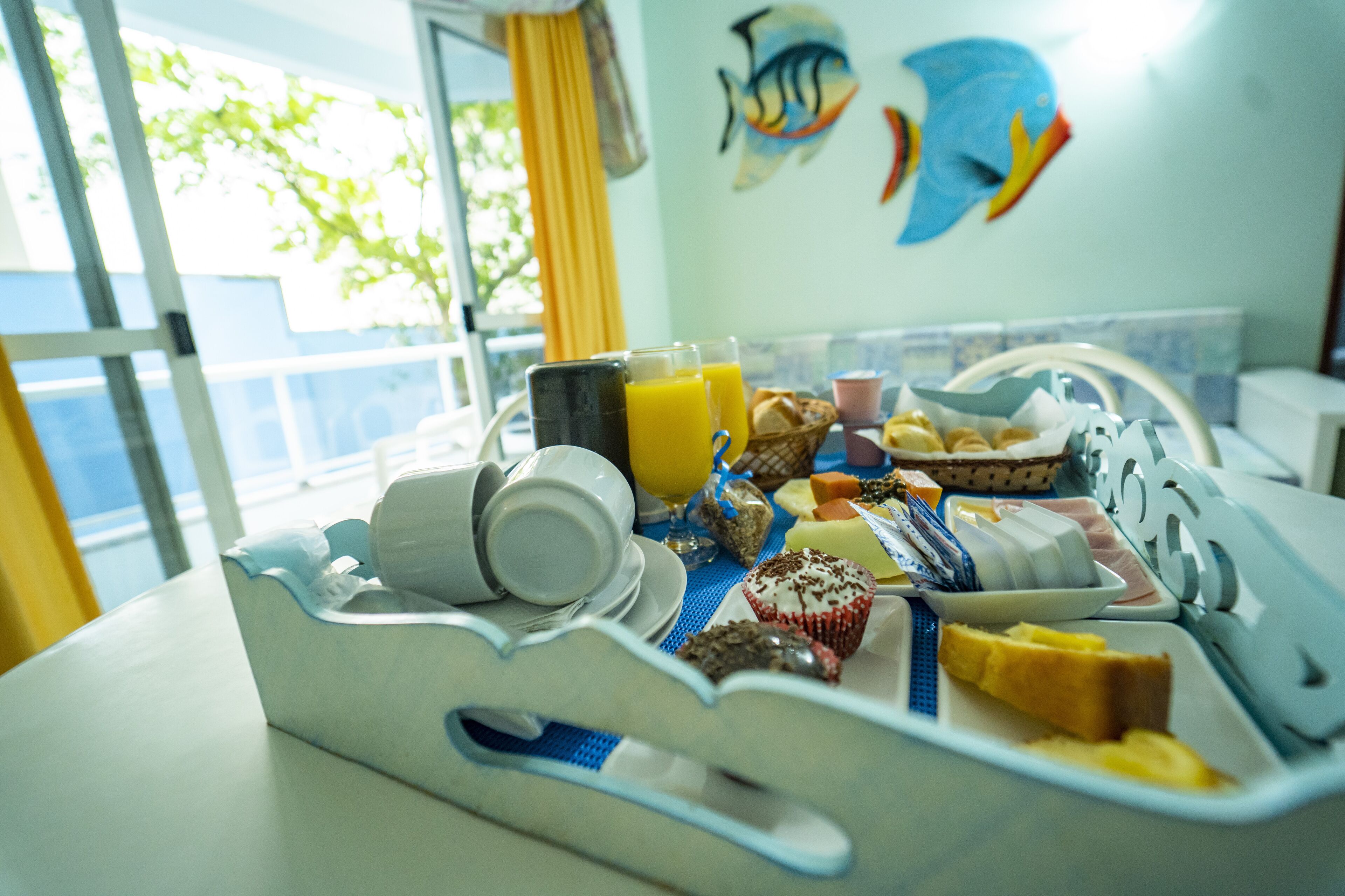 Una bandeja de desayuno con diferentes alimentos y un vaso de jugo de naranja sobre una mesa en una habitación con vistas al exterior.