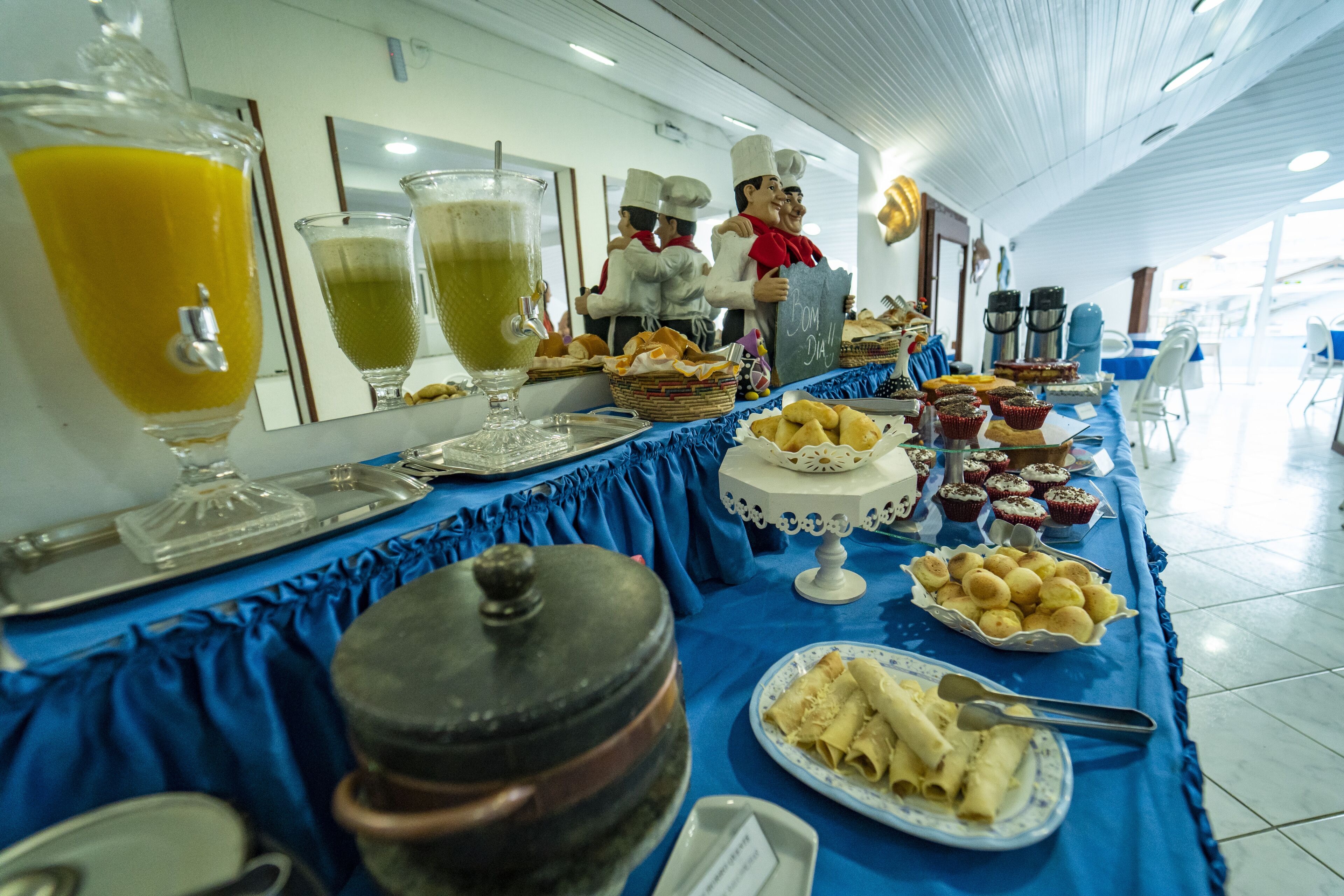 Une table de buffet proposant divers plats et boissons, notamment une grande carafe de jus d’orange, un verre de jus vert et un gâteau.