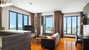 A modern living room with a sofa, a coffee table, and a TV. There are large windows with curtains, a wooden floor, and a view of the city. - Lumen Bangkok Srinakarin (Bangkok)